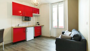 Cocina pequeña con armarios rojos, un microondas y un refrigerador. Hay una ventana con persianas y un sofá negro con cojines.