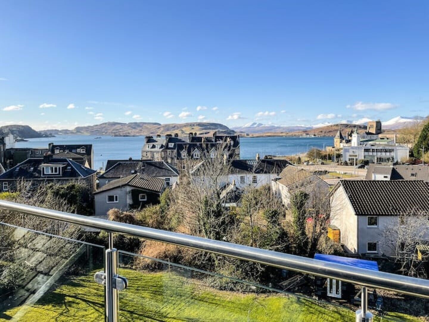 A coastal town with traditional houses, a clear blue sky, and a view of the sea and mountains in the distance.