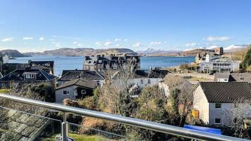 A coastal town with traditional houses, a clear blue sky, and a view of the sea and mountains in the distance.