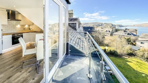 A modern kitchen with a dining area, large windows, and a view of a lake and town. - The Penthouse, Beautiful sea views, Centre of Oban (Oban)