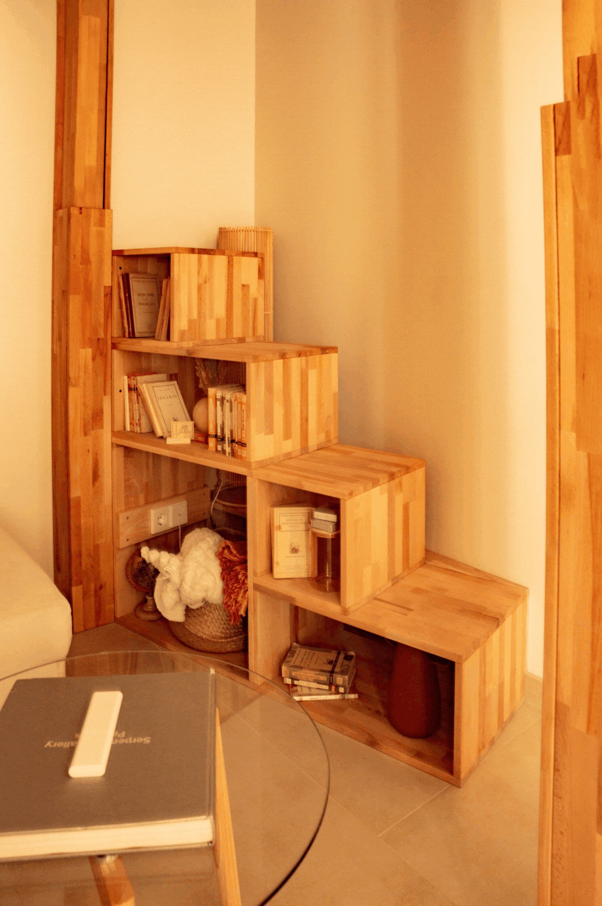 A corner of a room with a wooden bookshelf containing books and decorative items, a glass table with a book, and a white chair.