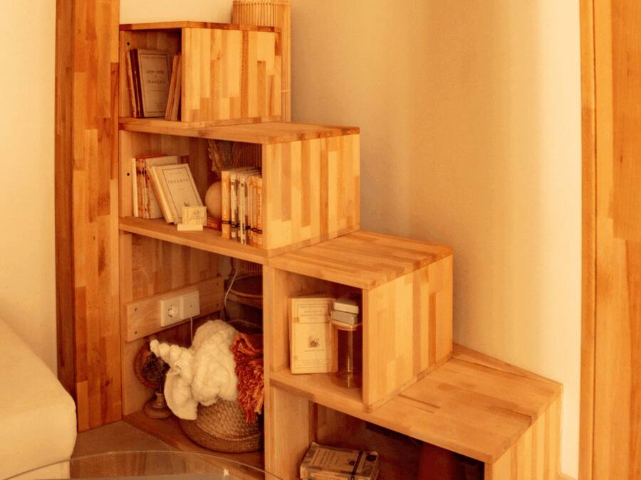 A corner of a room with a wooden bookshelf containing books and decorative items, a glass table with a book, and a white chair.