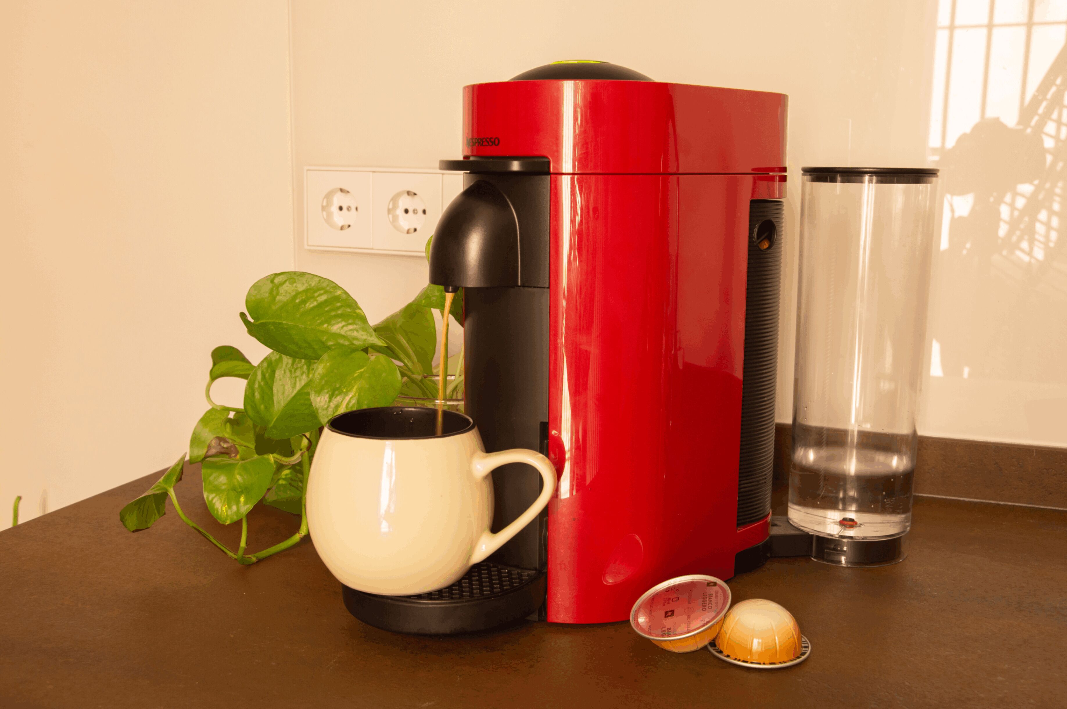 Een rode koffiemachine, een witte mok, een helder glas en een plant op een aanrecht.