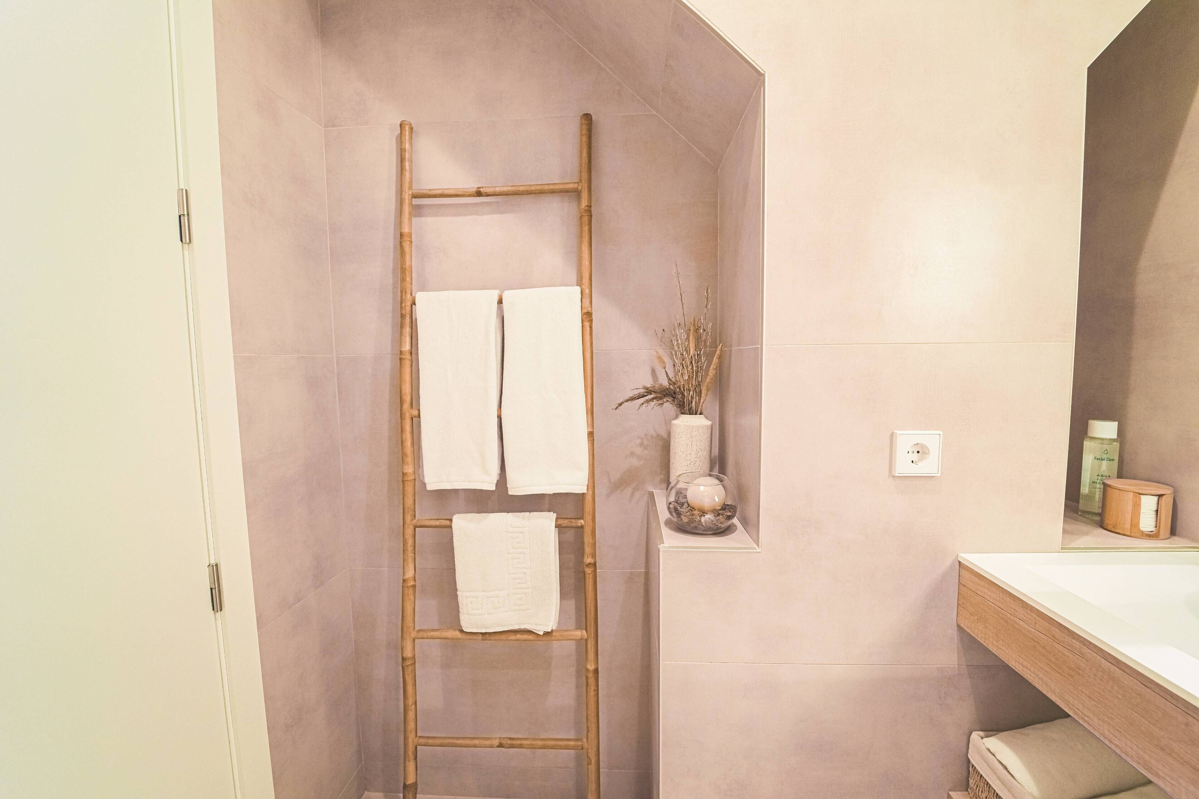 A bathroom with a wooden ladder, two white towels, a vase with dried plants, and a wooden cabinet with toiletries.