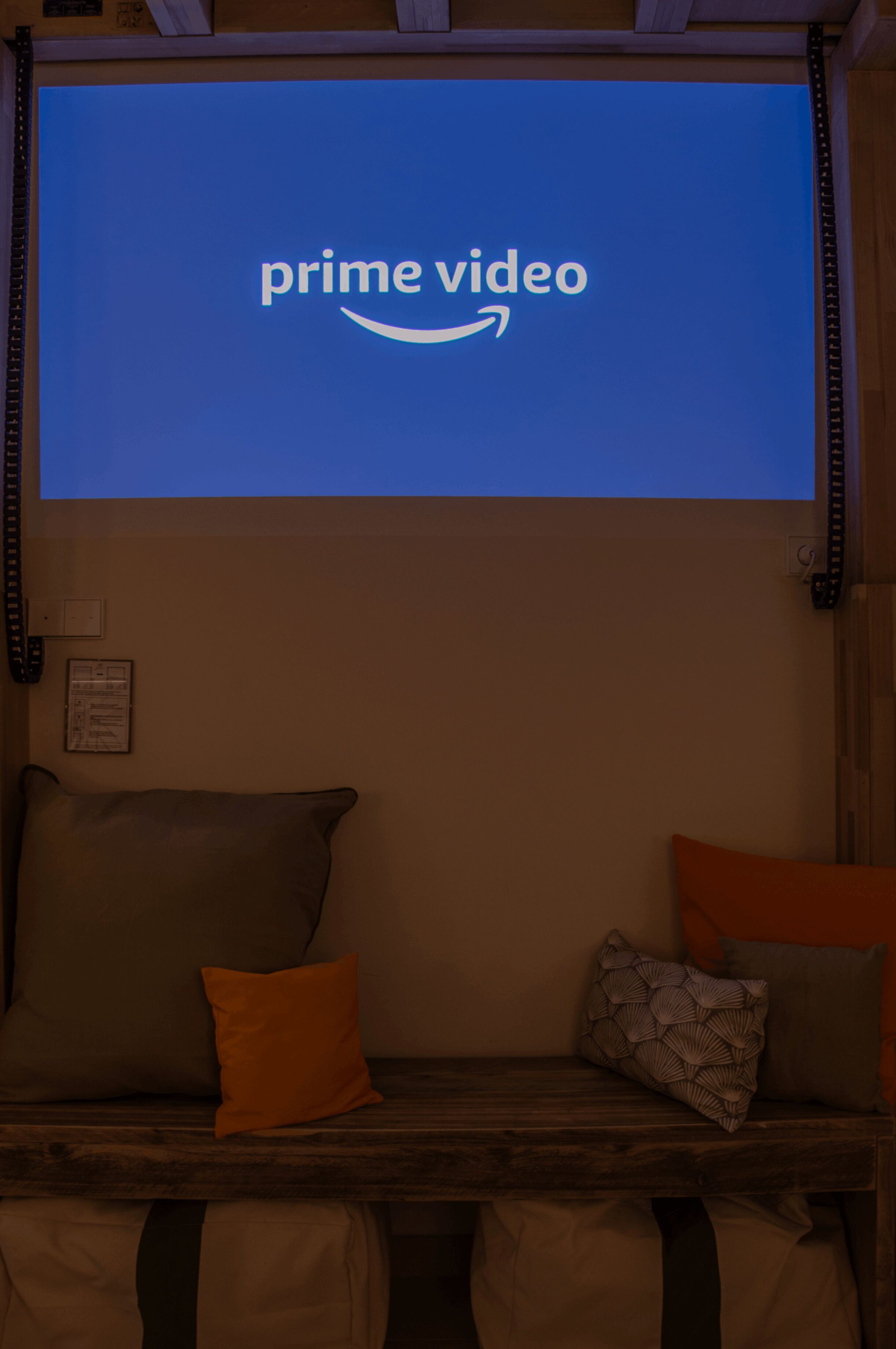Una habitación con una pantalla grande mostrando el logotipo de Prime Video, un banco de madera con cojines y un proyector instalado en la pared.