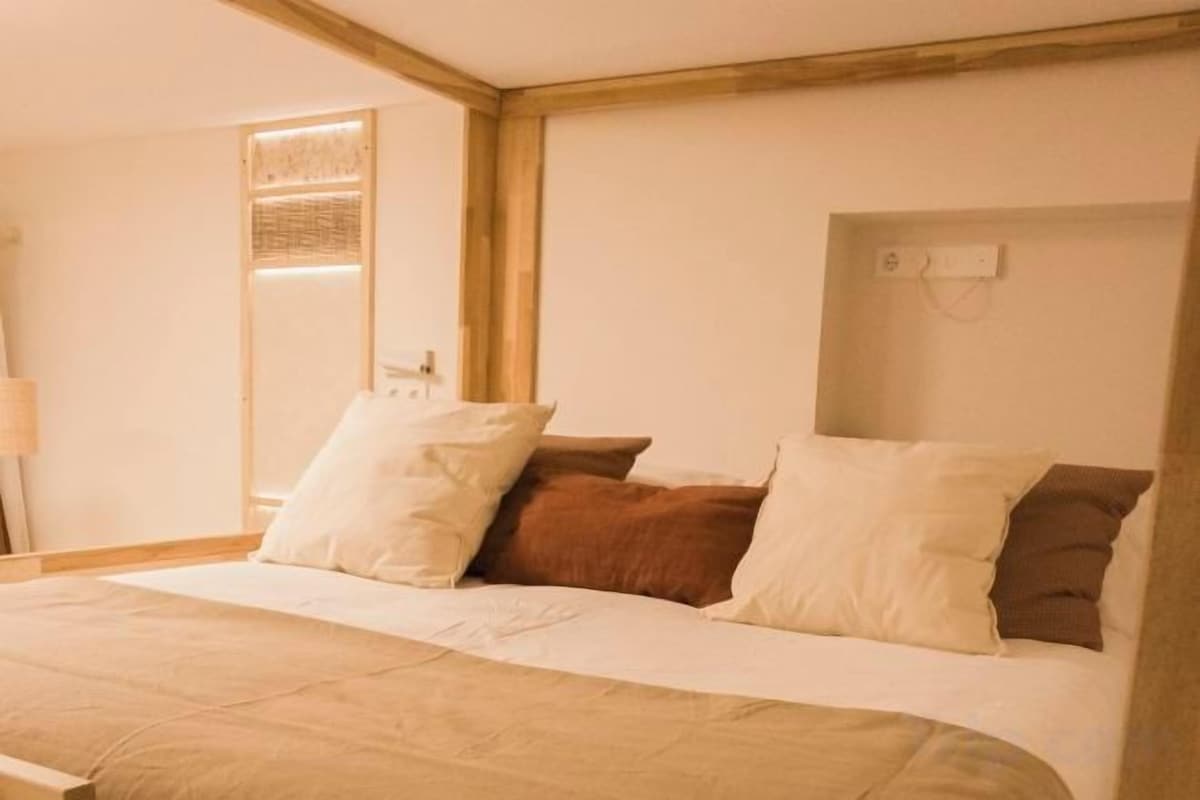 Een slaapkamer met een bed, kussens, een nachtkastje en een wandlamp.