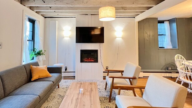 A modern living room with a fireplace, wooden ceiling beams, a flat-screen TV, and a dining area with chairs. - Historic house overlooking Rondout Creek (Kingston)