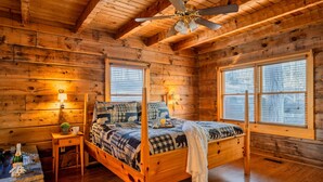 Un dormitorio de cabaña de madera con una cama con dosel, un ventilador de techo y ventanas amplias.