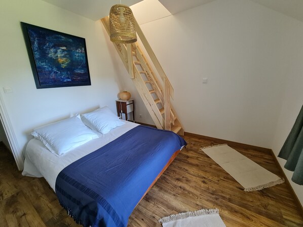A bedroom with a bed, a wooden ladder, a painting, and a small table with a lamp.