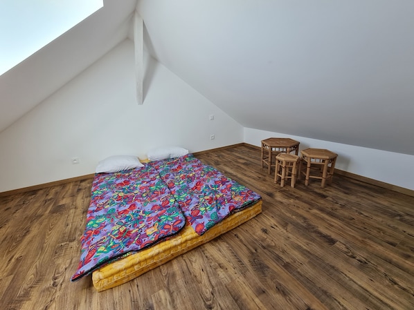 Een kleine kamer met een schuin dak, een bed, twee houten krukken en een raam.
