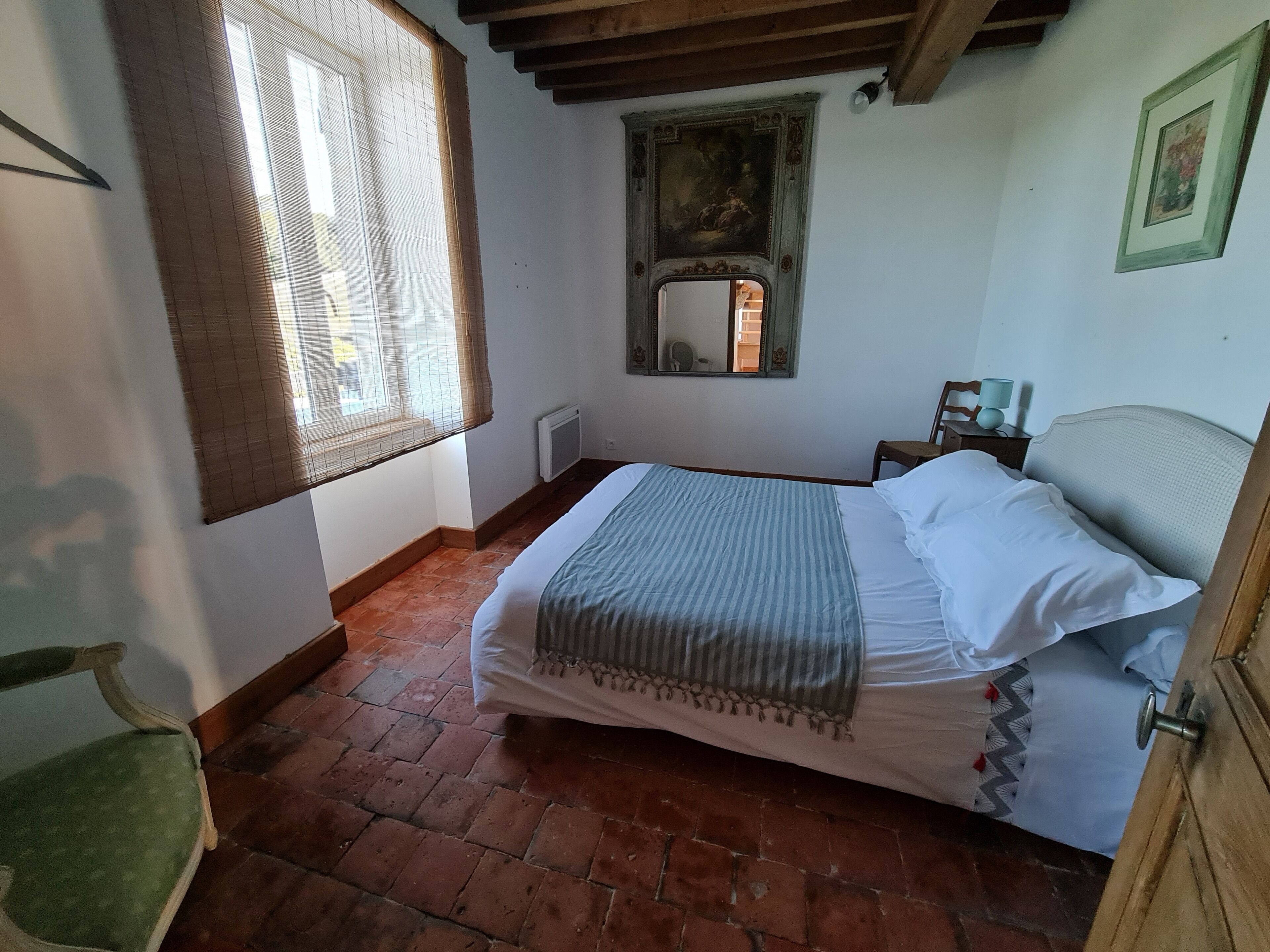 Um quarto com cama, uma cadeira, janela com persianas, radiador e espelho na parede.