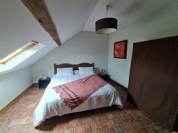 Une chambre avec un plafond en pente, un puits de lumière, un lit avec une literie blanche, une couverture marron, une table de chevet avec une lampe et un tableau rouge accroché au mur.