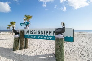 Un panneau en bois portant l’inscription « MISSISSIPPI GULF COAST » et un panneau « bienvenue » en haut, le tout entouré de palmiers sur une plage de sable.