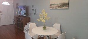 A dining area with a round table and four chairs, a television on a wooden cabinet, decorative wall art, and a door with a semi-circular window. - Oak Shores 157 | Just a Short Walk to the Beach! (Biloxi)