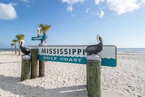 Un letrero de madera con la inscripción 'MISSISSIPPI GULF COAST' y un letrero de 'welcome' encima, rodeado de palmeras sobre una playa arenosa.