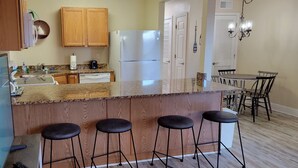A kitchen with wooden cabinets, a white refrigerator, a countertop with a coffee maker, and a dining area with a table and chairs. - Oak Shores 155 | First Floor Unit, Just a Short Walk to the Beach! (Biloxi)