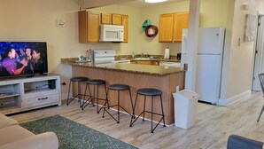 A compact living space with a kitchen area featuring wooden cabinets, a microwave, a refrigerator, and a bar stool setup. - Oak Shores 155 | First Floor Unit, Just a Short Walk to the Beach! (Biloxi)