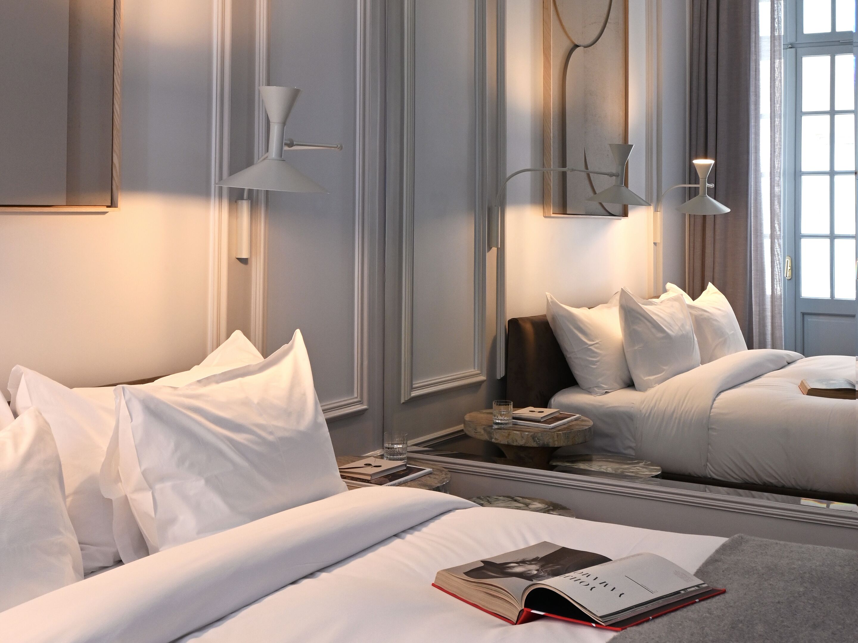 A hotel room with a neatly made bed, a bedside table with a book and glass, and a large mirror reflecting the room.