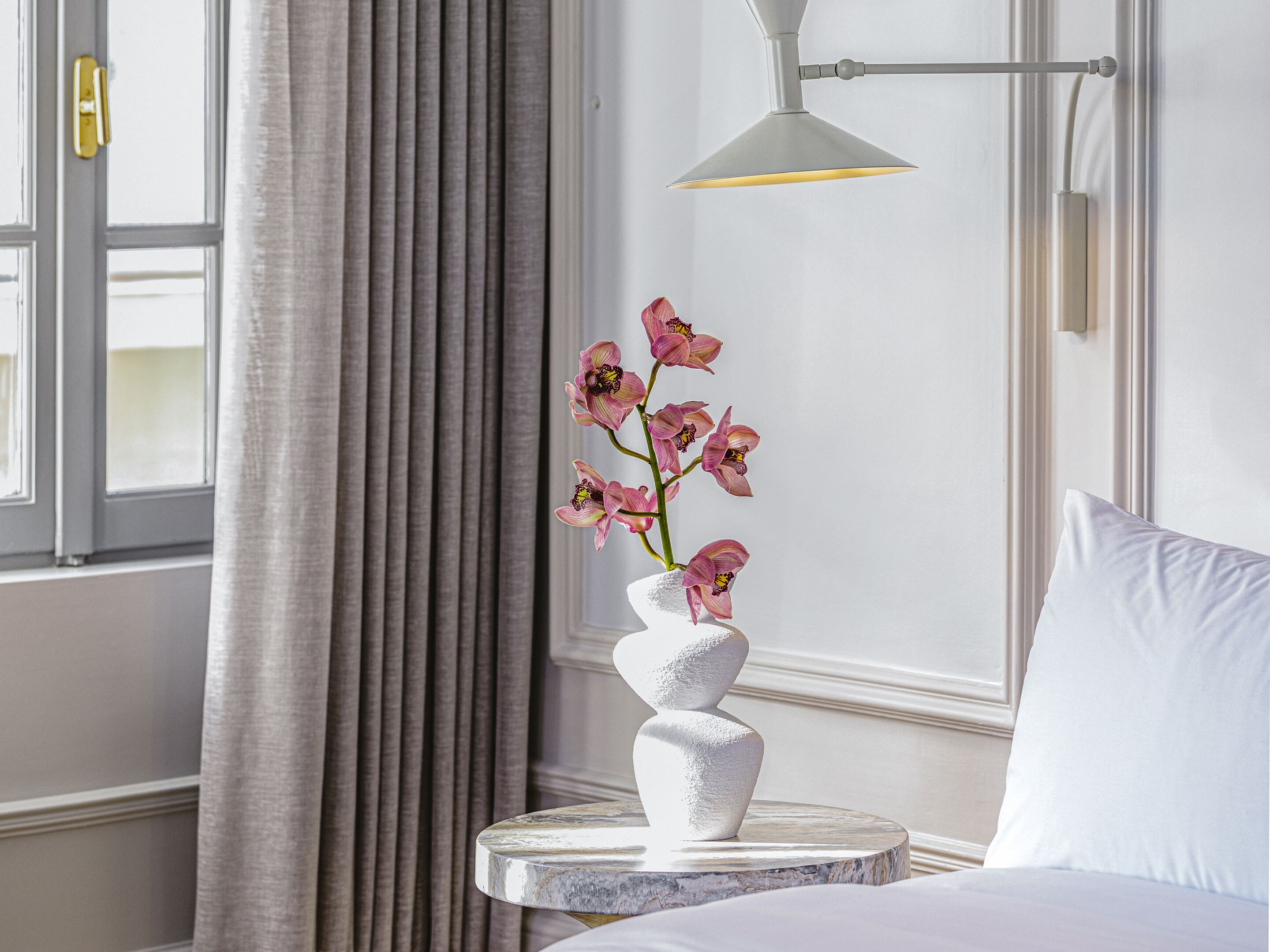 Quarto de hotel com uma cama, um vaso branco com flores cor-de-rosa, uma mesa redonda e uma luminária pendente moderna.
