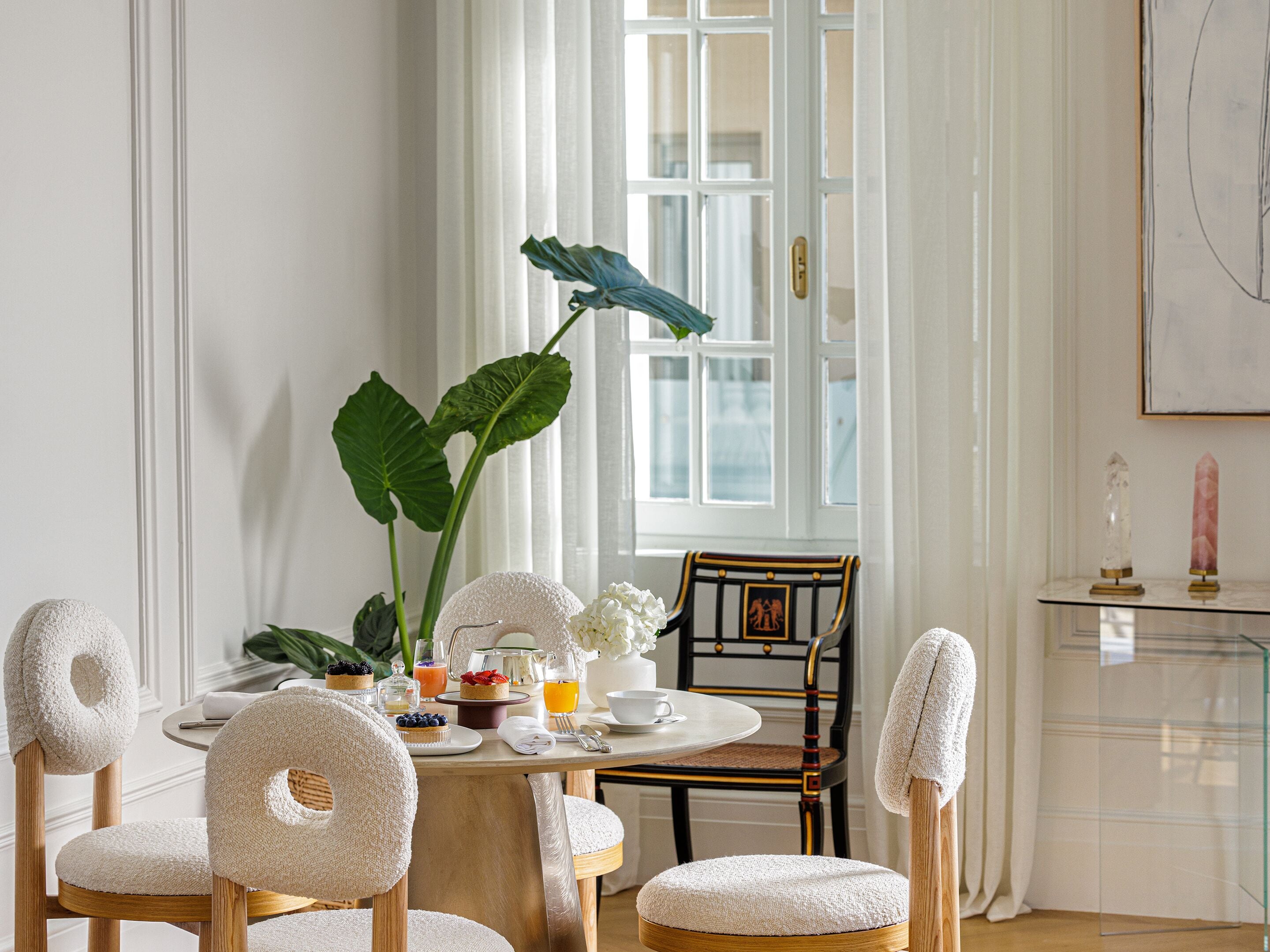 A dining area with a round table, chairs, a plant, and a window with curtains.