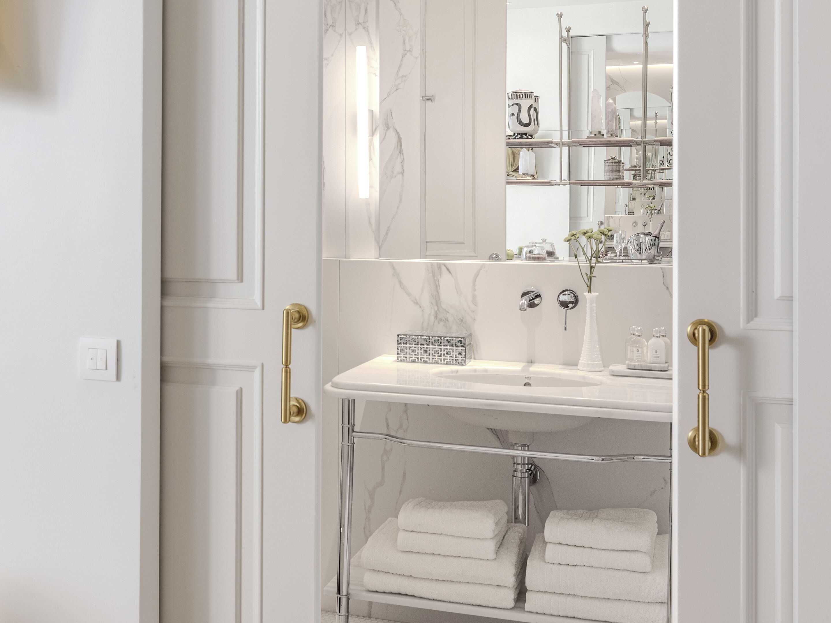A bathroom with a white marble sink, a glass shelf with towels, and a mirror reflecting the room.