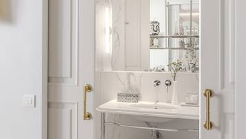 Une salle de bain avec un lavabo en marbre blanc, une étagère en verre où sont posées des serviettes et un miroir qui reflète la pièce.