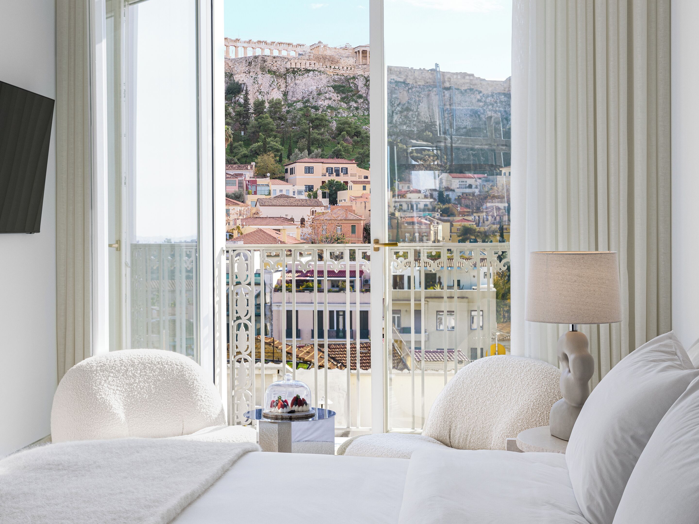 Habitación de hotel con balcón, cama con ropa de cama blanca, lámpara y vista a un paisaje urbano con edificios y una montaña.