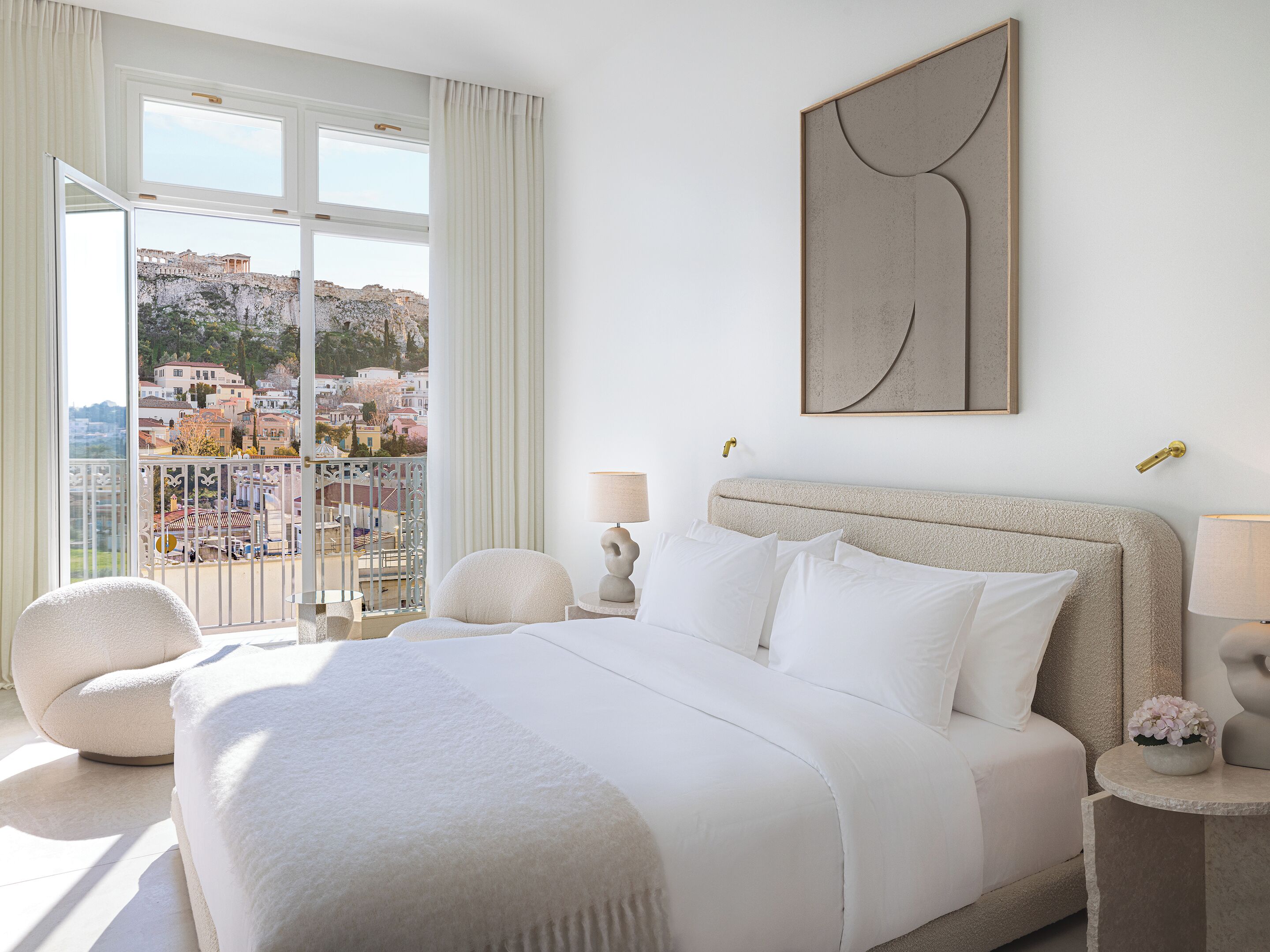 A hotel room with a bed, a view of a cityscape, a balcony, and a modern abstract painting.