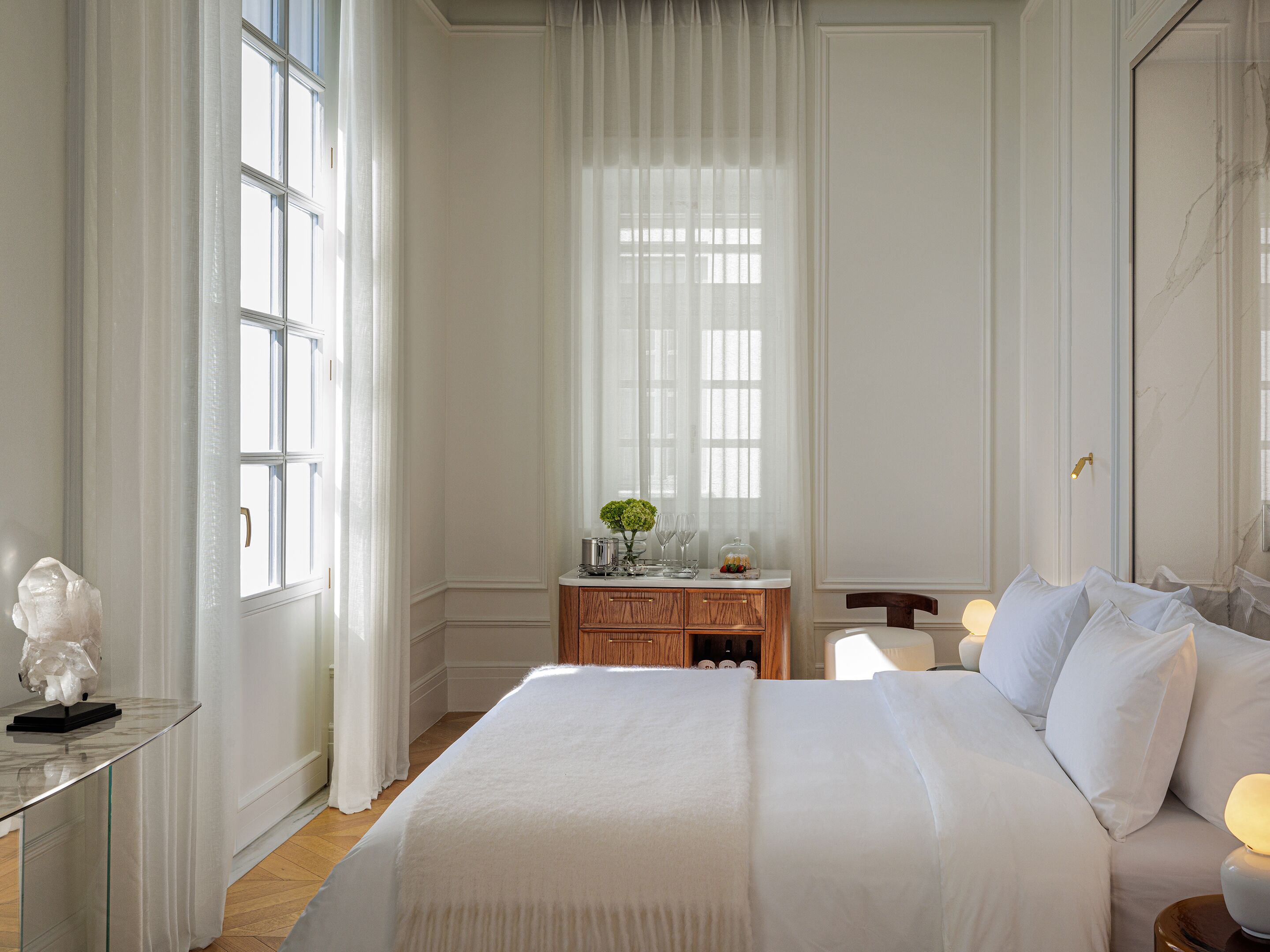 Quarto de hotel com uma cama grande, mesa de cabeceira, cômoda e janela com cortinas leves.