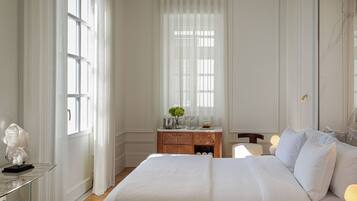 Quarto de hotel com uma cama grande, mesa de cabeceira, cômoda e janela com cortinas leves.