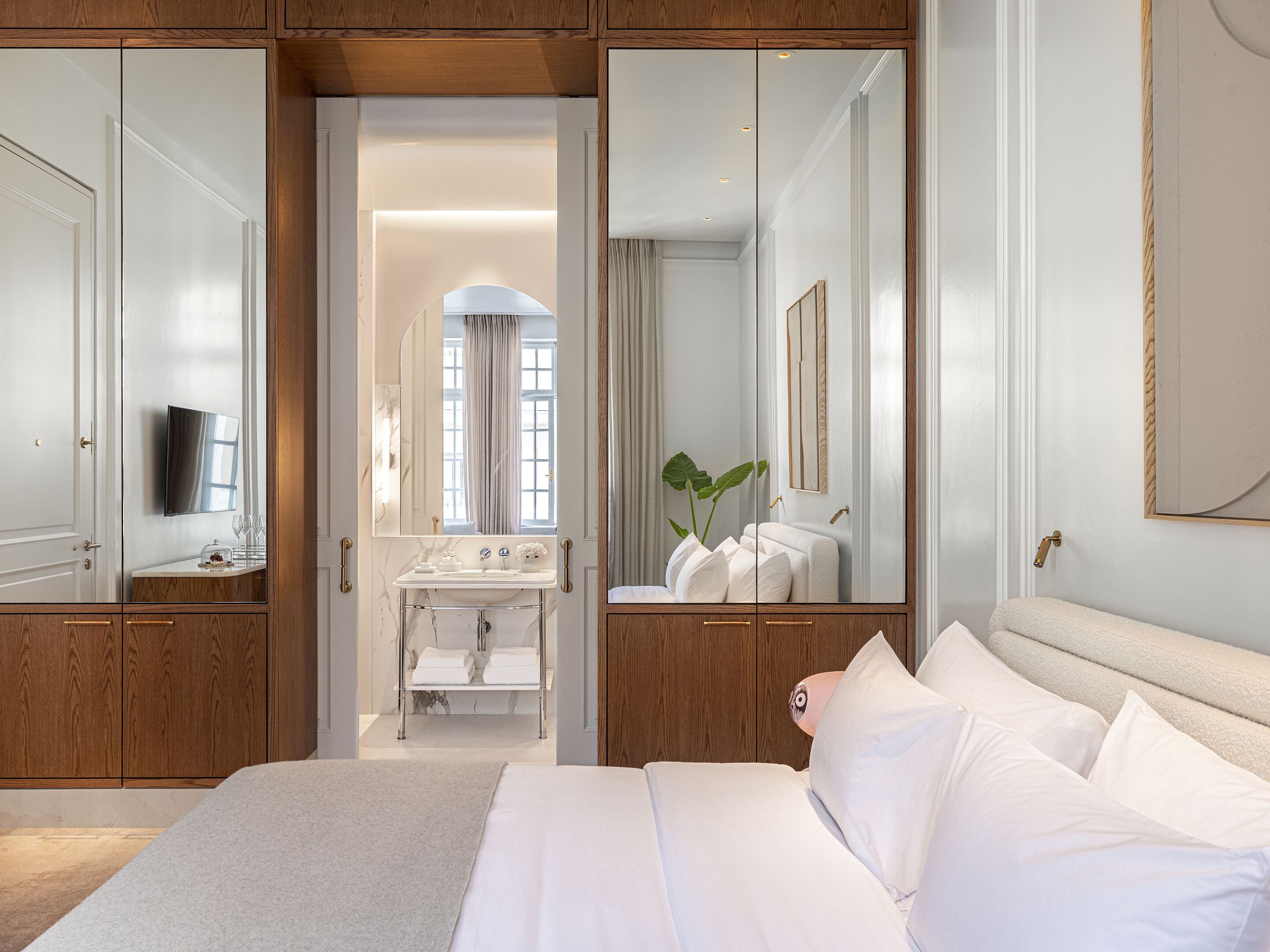 A bedroom with a large bed, a bedside table, a mirror, a television, and a bathroom with a sink and towel rack.