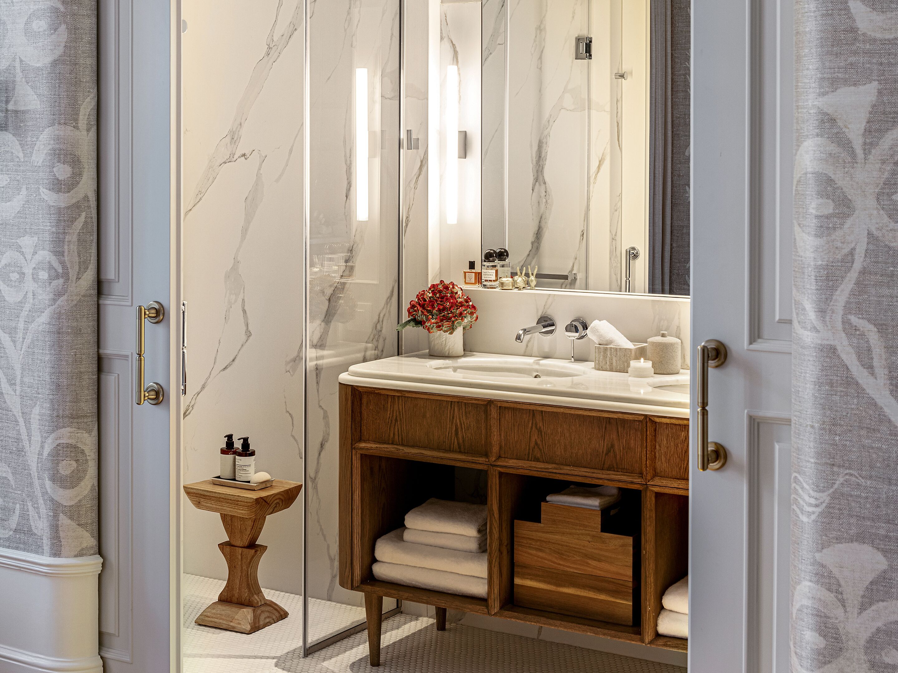 A bathroom with a marble wall, wooden vanity, and a small wooden stool with toiletries.