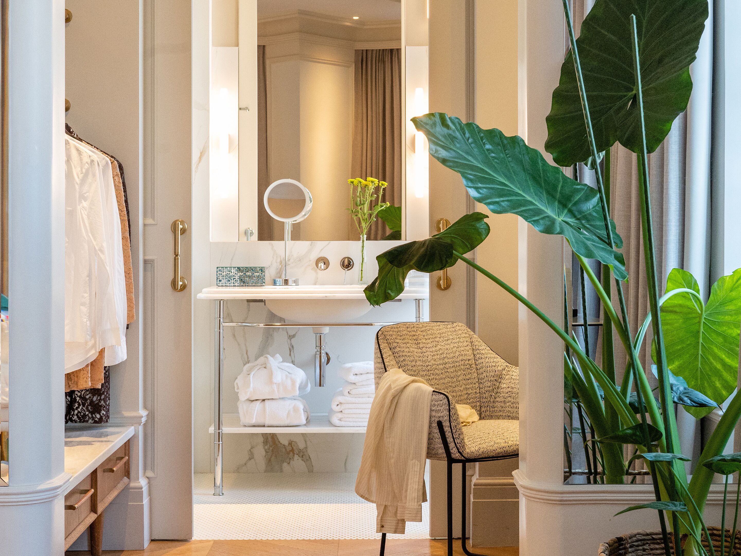 A bathroom with a marble sink, a round mirror, a towel rack, and a chair.
