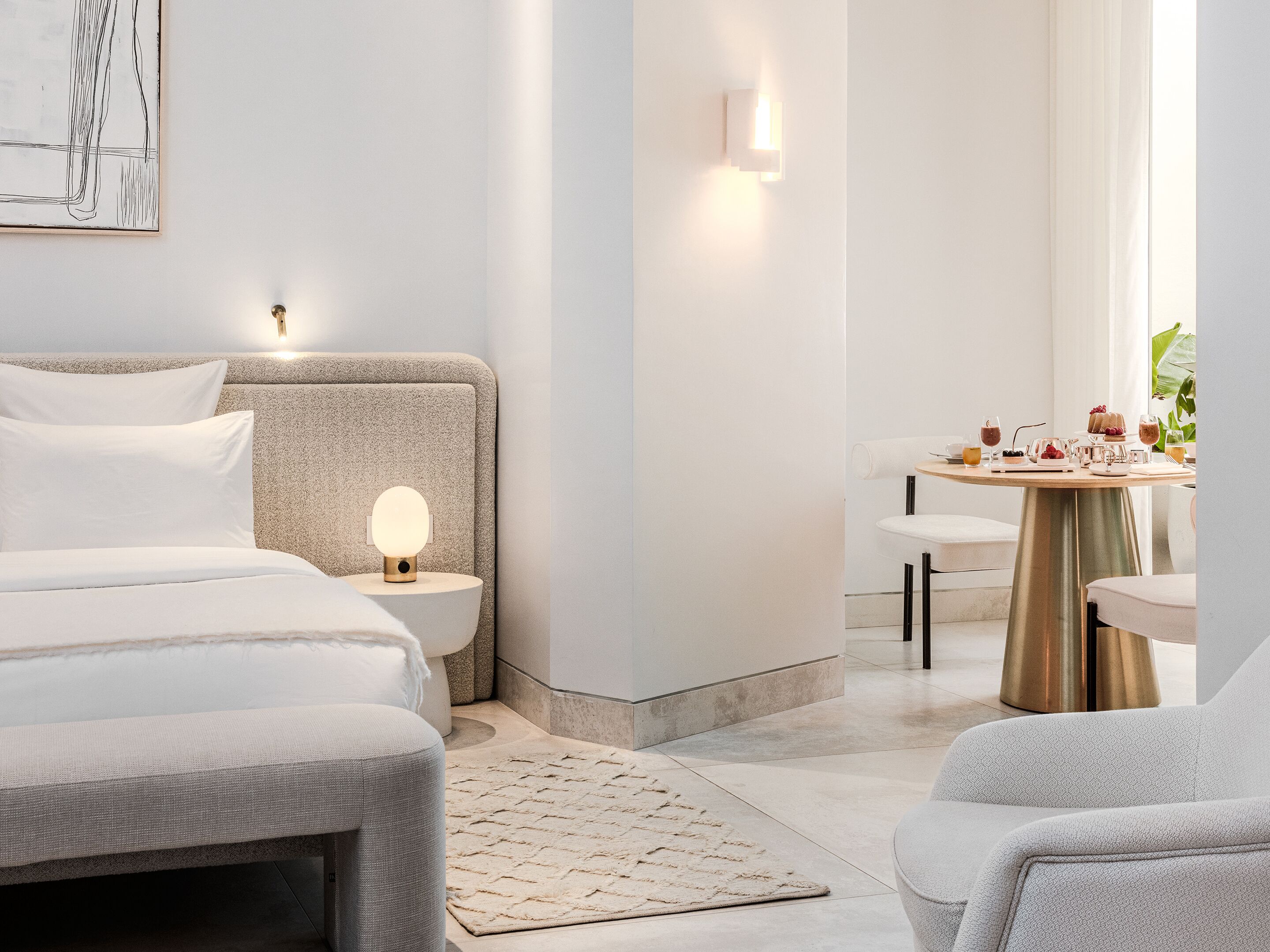A modern hotel room with a bed, bedside table with a lamp, a round dining table with chairs, and a view of the outdoors.
