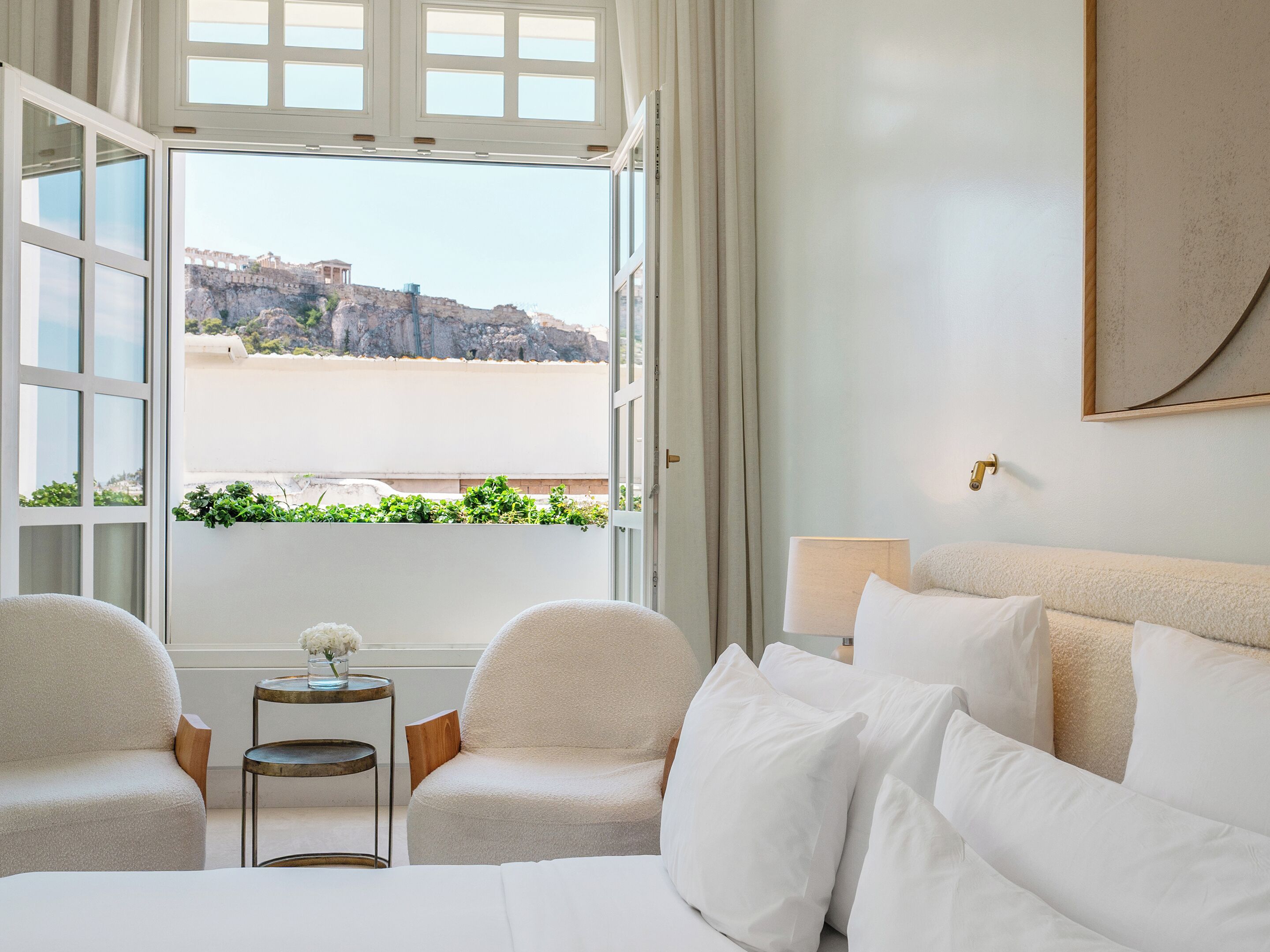 A hotel room with a bed, two chairs, a small table with a vase, and a view of a beach and rocky cliffs.