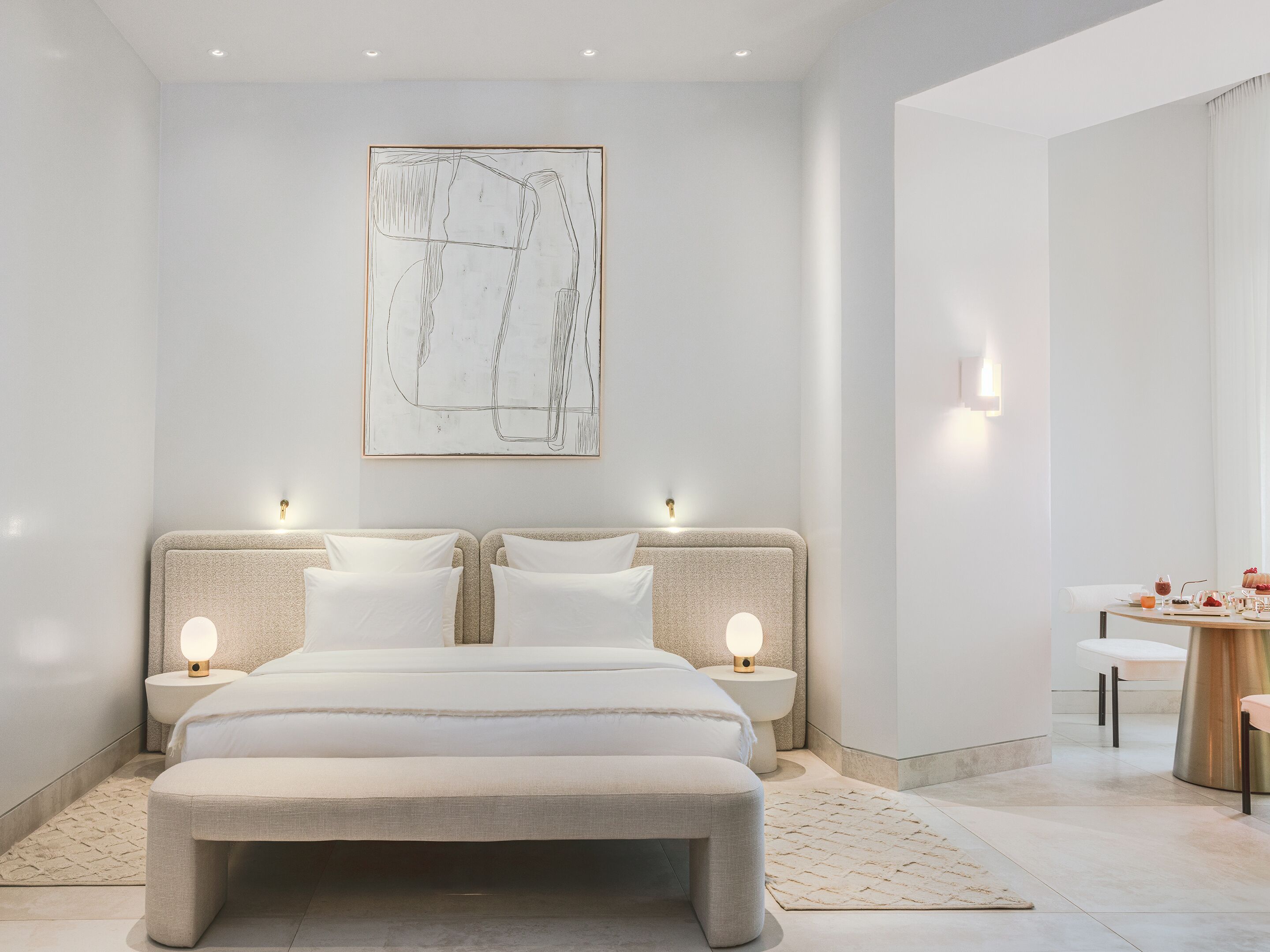Un dormitorio moderno con una cama grande, mesitas de noche, una obra de arte abstracta en la pared y un rincón de desayuno con mesa y sillas.