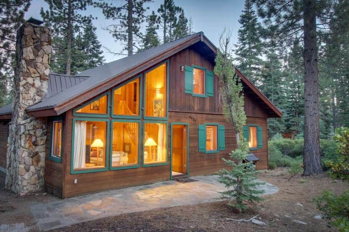A wooden cabin with large windows, a stone chimney, and a stone foundation.