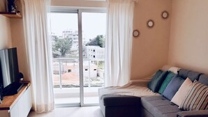 A modern living room with a grey sofa, a flat-screen TV on a wooden stand, and a sliding glass door leading to a balcony.