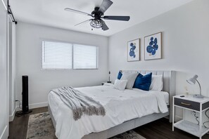 A bedroom with a bed, a bedside table, a ceiling fan, a window with blinds, and two framed artworks on the wall.