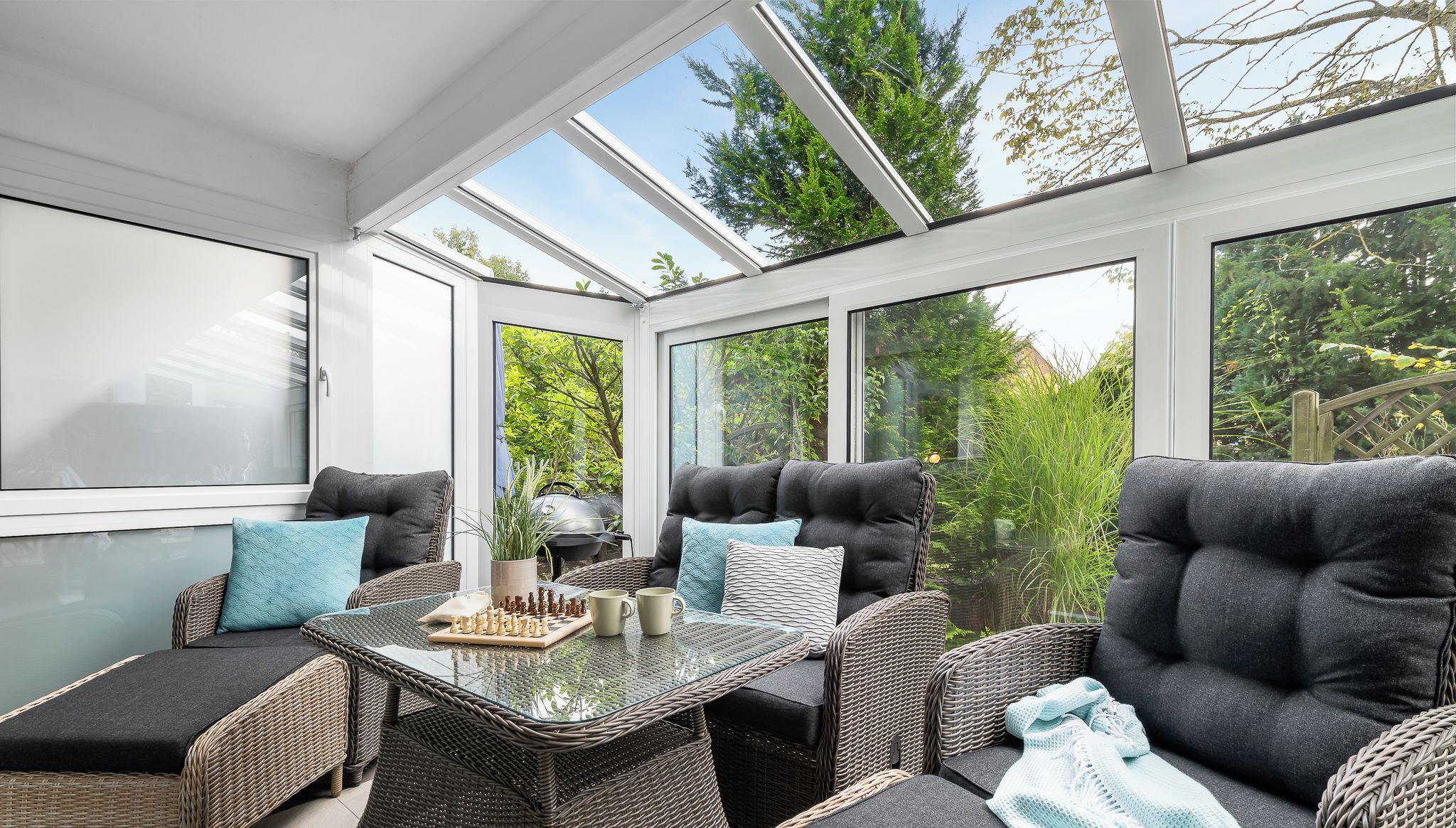 A sunroom with wicker furniture, a glass table, and a view of greenery.