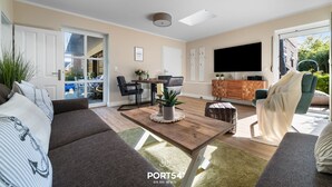 A modern living room with a sofa, coffee table, and TV. There is a dining area with chairs and a table. The room has large windows and a ceiling light. - Ferienwohnung Deck 3 Büsum (Büsum)