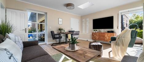 A modern living room with a sofa, coffee table, and TV. There is a dining area with chairs and a table. The room has large windows and a ceiling light.