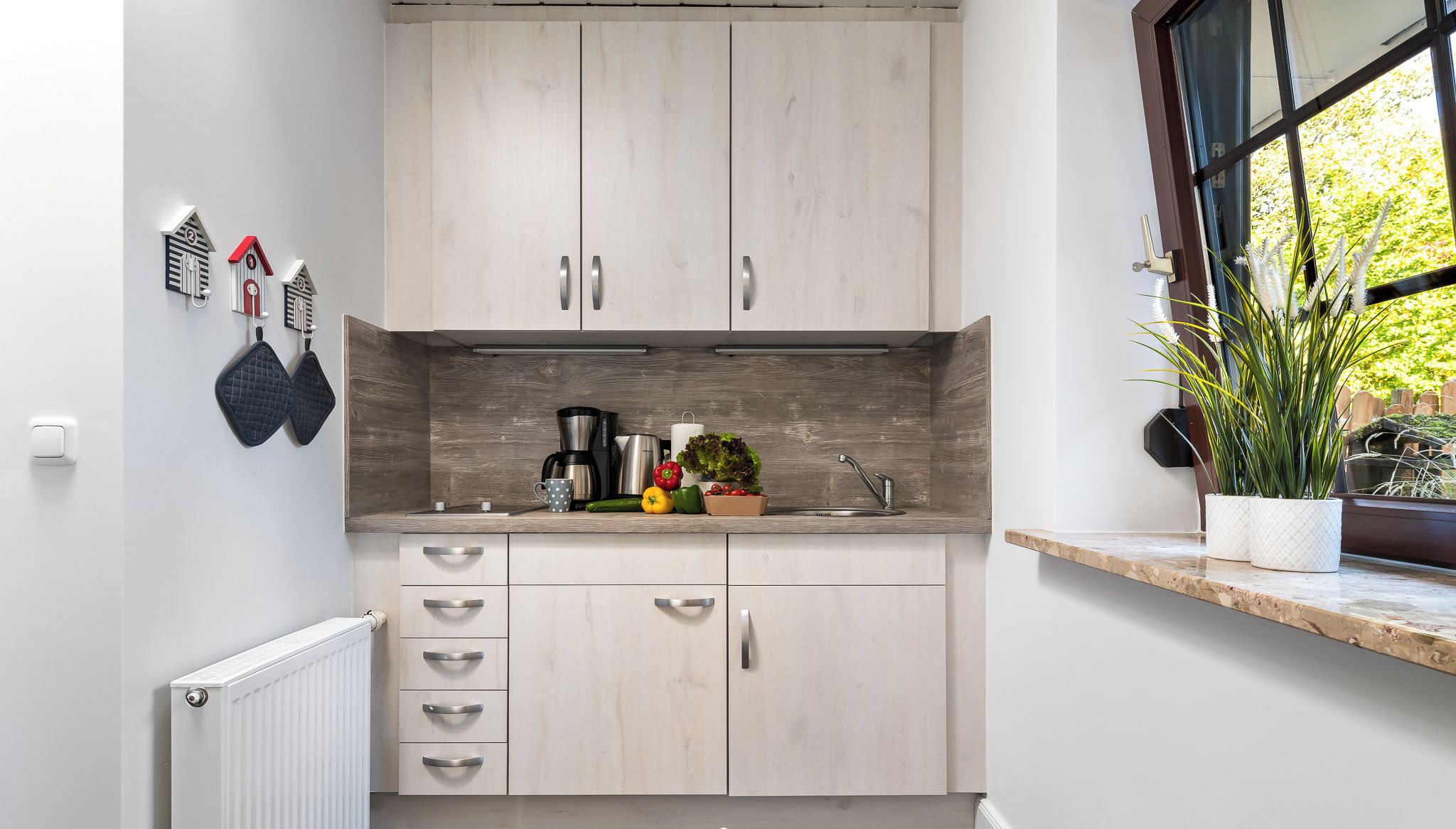 A modern kitchen with light wood cabinets, a built-in oven, and a window with a view of greenery.