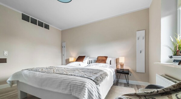 Une chambre moderne avec un grand lit, des lampes de chevet, une petite table et une fenêtre ornée de plantes.