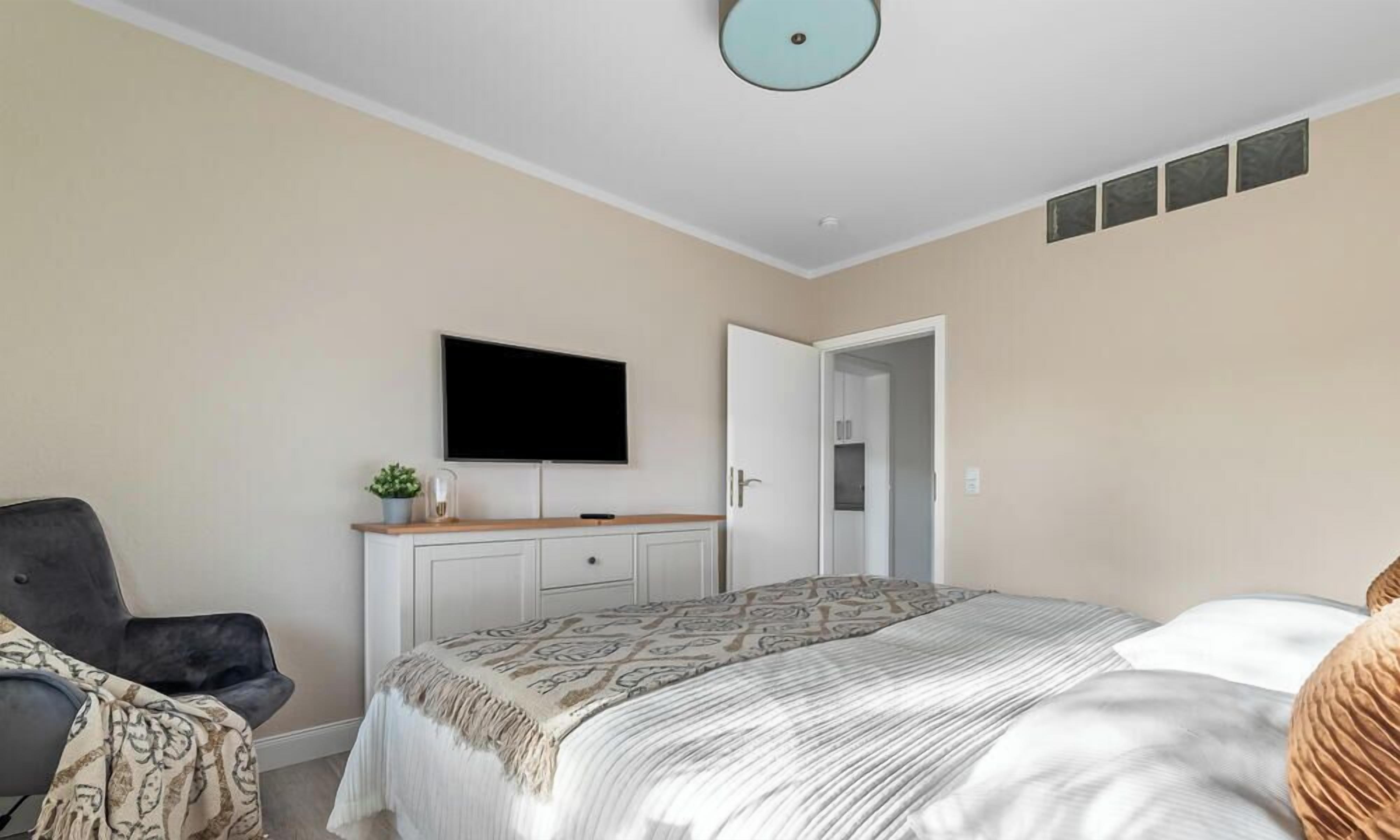 A bedroom with a bed, a TV on the wall, a chair, and a door leading to another room.