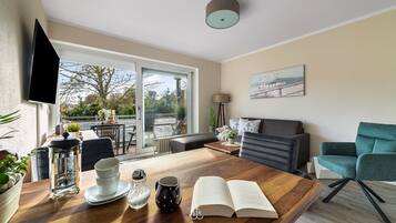 A modern living room with a wooden coffee table, a sofa, and a teal armchair. There is a large window offering a view of the outdoors, and a flat-screen TV mounted on the wall.