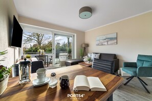 A modern living room with a wooden coffee table, a sofa, and a teal armchair. There is a large window offering a view of the outdoors, and a flat-screen TV mounted on the wall.