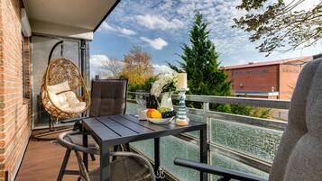 A balcony with wicker furniture, a table with fruits and a candle, and a view of trees and buildings.