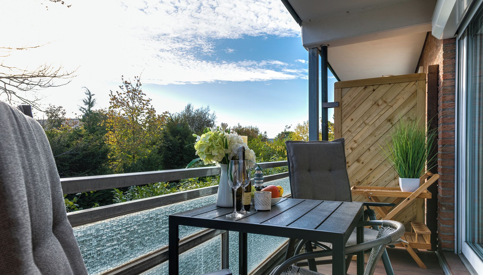 Een balkon met een tafel gedekt voor twee personen, een bloempot en een fles wijn.