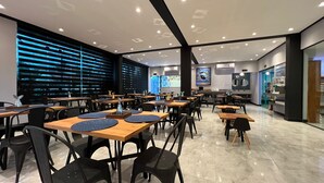 A modern restaurant with multiple tables and chairs, a kitchen area, and a large window with blinds. - Hotel la serena (Sao Sebastiao)